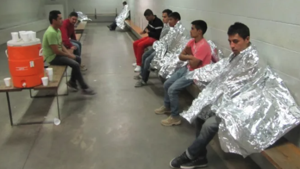 eight migrant men sitting in a concrete and cinderblock holding cell, some wearing space blankets